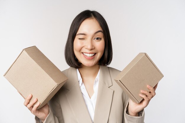 Image of saleswoman asian businesswoman holding boxes with company brand product smiling at camera standing against white background