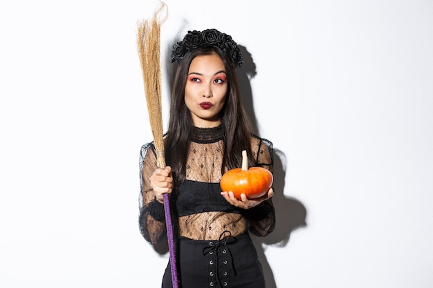Image of sassy witch in gothic lace dress, holding broom and pumpkin, looking at upper left corner with halloween banner, white background.