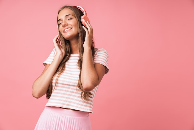 Image of satisfied beautiful woman wearing striped clothes smiling and listening to music with headphones isolated over pink wall