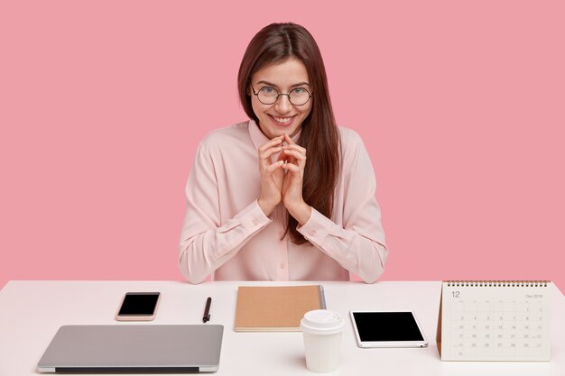 Image of satisfied European lady office worker being real perfectionist, keeps hands together, smiles gently