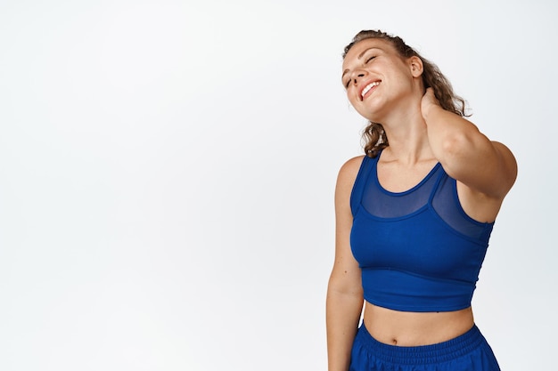 Изображение довольной спортивной девушки, потирающей шею и улыбающейся. Фитнес-женщина чувствует удовольствие после продуктивной тренировки, выполнения упражнений, на белом фоне