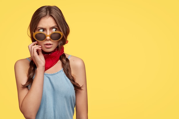 Изображение испуганной европейской женщины с прямыми волосами, в модных солнцезащитных очках, красной банадой на шее, смотрит с мрачным выражением лица в сторону, показывает свободное место на желтой стене для текста