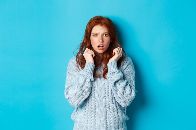 Image of scared teenage girl with red hair, jumping startled and looking alarmed, standing over blue background