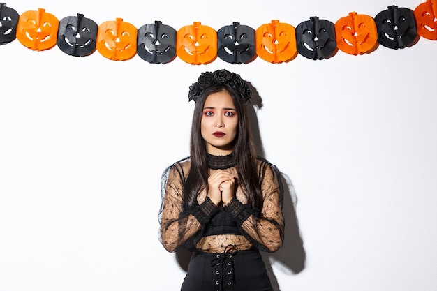 Free photo image of scared and worried asian woman in witch costume looking concerned, wearing witch costume and standing against pumpkin banners.