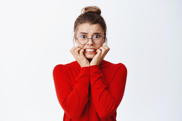 Image of scared young female employee in glasses panicking, biting finger nails from fear and anxiety, trembling of something scary, standing on white