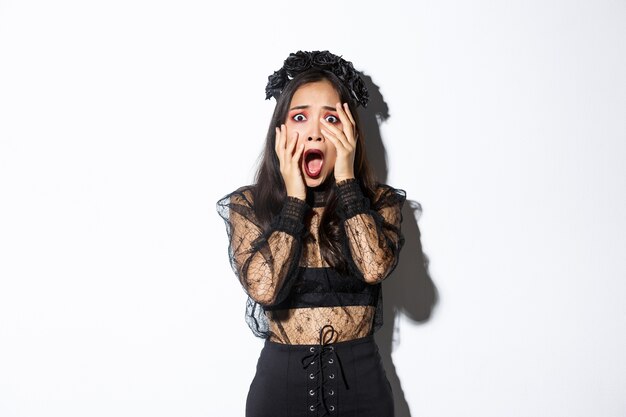 Image of scared young woman screaming and looking frightened at something creepy on halloween, wearing gothic dress and wreath, holding hands on face and trembling from fear.