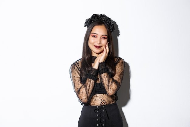 Image of sensual smiling asian gothic girl, woman in halloween costume gently touching face and looking at camera dreamy, standing over white background.