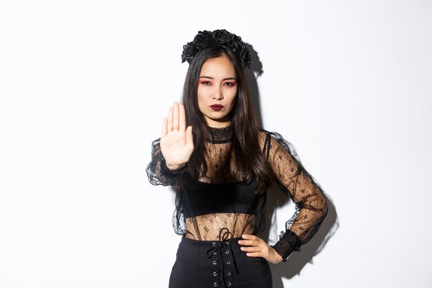 Image of serious asian woman in halloween costume of witch, showing stop gesture