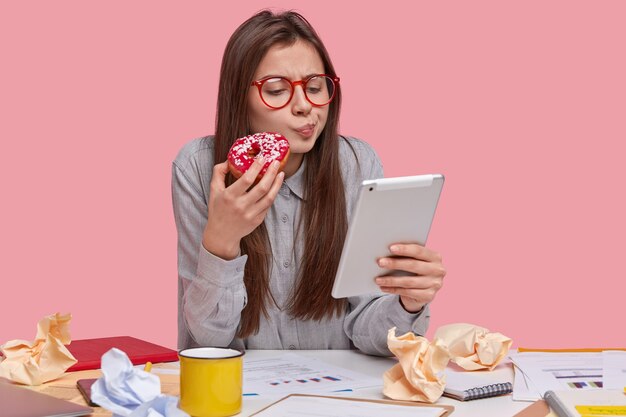 Image of serious beautiful lady holds delicious doughnut, watches training video on touchpad, prepares business report, studies graphic