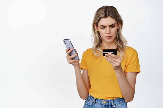 Image of serious blond girl holding mobile phone looking at credit card number with concentrated face trying to pay online standing over white background