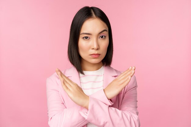 Image of serious corporate woman asian businesswoman showing stop cross gesture disapprove smth standing in suit over pink background