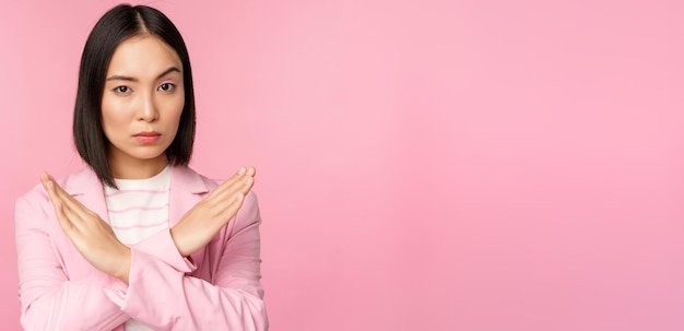 Image of serious corporate woman asian businesswoman showing stop cross gesture disapprove smth standing in suit over pink background