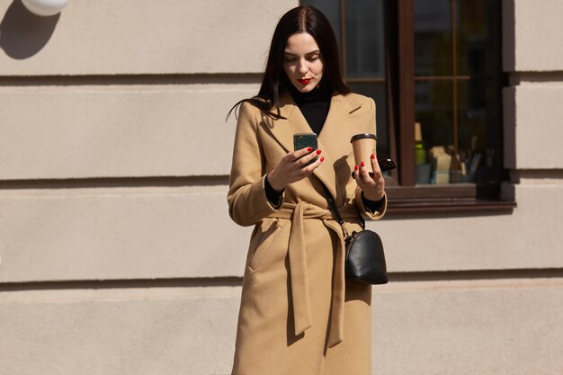 Образ серьезной молодой женщины в элегантном бежевом пальто, используя ее телефон на улице солнечного города
