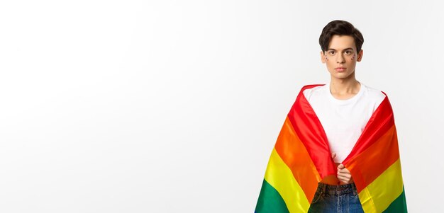 Изображение серьезно выглядящего гендерно-изменчивого человека с блестками на лице, стоящего с радужным флагом лгбтк