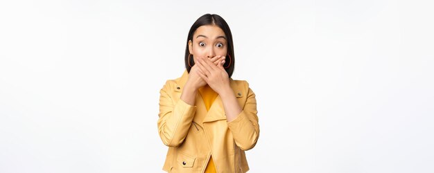 Image of shocked asian girl shuts mouth close lips and looking speechless startled face expression standing over white background