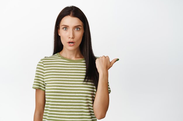 Image of shocked brunette woman pointing right looking amazed at camera showing smth amazing standing over white background