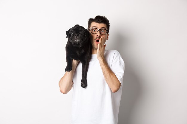 Image of shocked dog owner staring at camera and gasping impressed, cute black pug sitting on his shoulder and staring at you, white background.