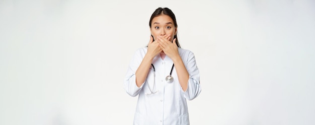 충격을 받은 아시아 여성 의사의 이미지는 손으로 입을 막고 배경 위에 서 있는 걱정스러운 금기 제스처를 보고 입술을 가리고 있습니다.
