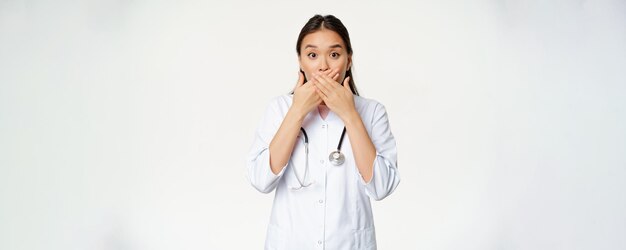 ショックを受けた女性アジア人医師の画像は、手を閉じて口を閉じ、白い背景の上に立っている心配しているタブージェスチャーを見て彼女の唇を覆っています