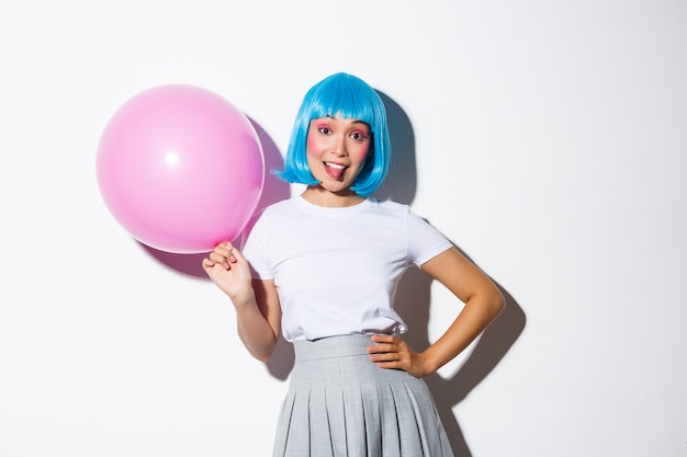 Image of silly party girl in blue wig celebrating holiday, holding pink balloon and showing tongue, standing backgound.