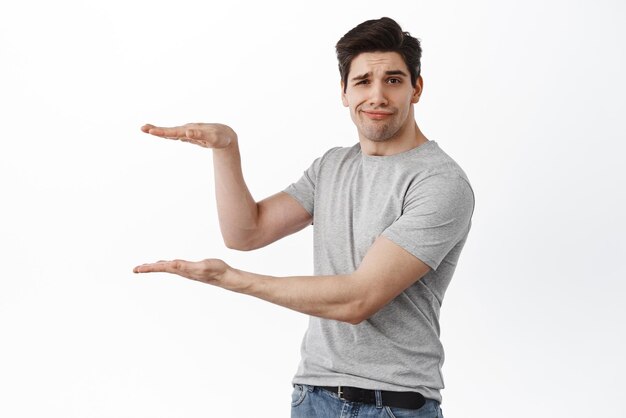 Image of skeptical brunette guy holding empty copy space between hands showing hand box gesture as if display product or logo looking displeased or unamused white background