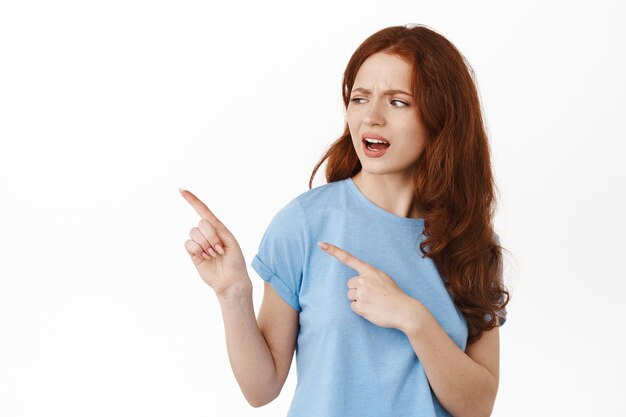 Image of skeptical and confused redhead girl, pointing and looking left at something strange, dont like promotion, having douts in advertisement, standing in t-shirt against white background.