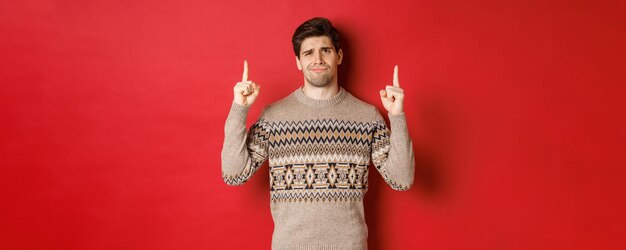 Image of skeptical and unamused handsome guy, showing bad christmas offer, pointing fingers up and grimacing displeased, express dislike, standing over red background