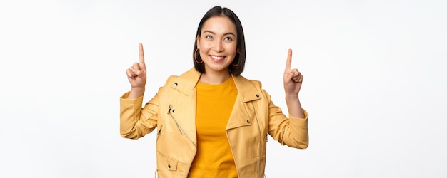 Immagine di una donna bruna asiatica sorridente che punta il dito verso l'alto mostrando pubblicità con la faccia felice in posa su sfondo bianco