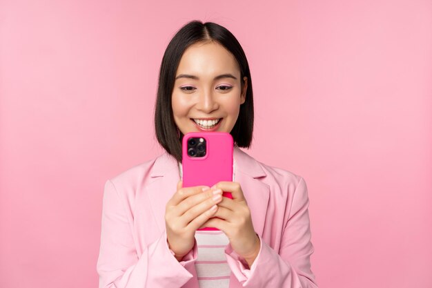 Image of smiling asian corporate woman in suit looking watching on smartphone app using mobile phone application standing over pink background
