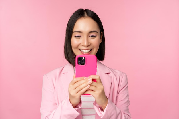 분홍색 배경 위에 서 있는 휴대폰 애플리케이션을 사용하여 스마트폰 앱을 보고 있는 정장 차림의 웃는 아시아 기업 여성의 이미지