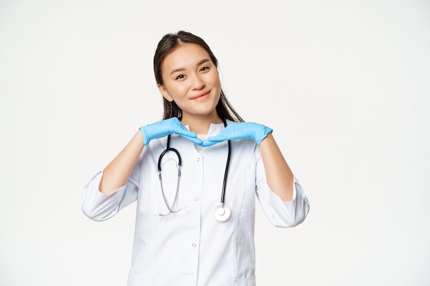Free Photo image of smiling asian doctor, dermatologist shows healthy skin and posing cute at camera, standing in medical uniform over white background.