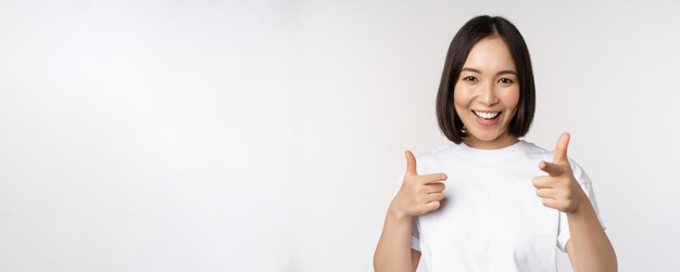 白い背景の上にTシャツで立っていることを祝福するように招待を選択してカメラに指を指している笑顔のアジアの女の子の画像
