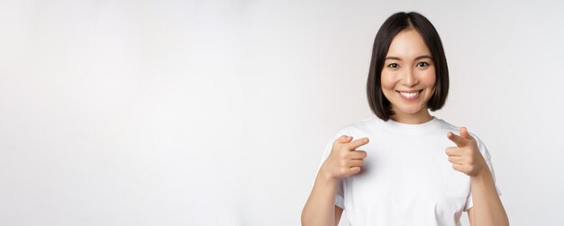 Image of smiling asian girl pointing fingers at camera choosing inviting you congratulating standing in tshirt over white background