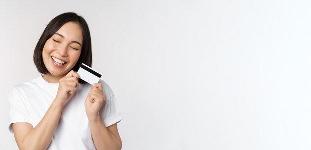 Image of smiling asian woman hugging credit card buying contactless standing in white tshirt over wh