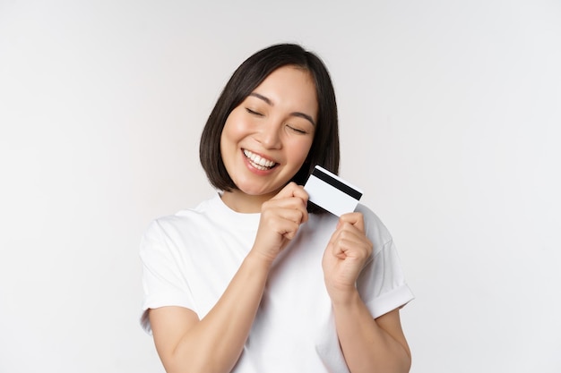 Image of smiling asian woman hugging credit card buying contactless standing in white tshirt over white background Copy space