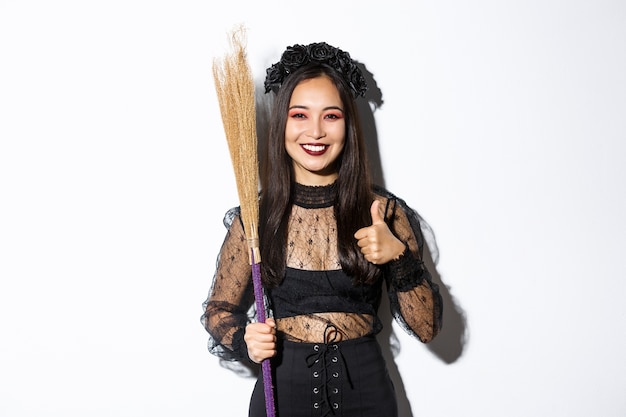 Изображение улыбающейся азиатской женщины в костюме ведьмы с метлой, показывая большие пальцы руки в одобрении, стоя на белом фоне.