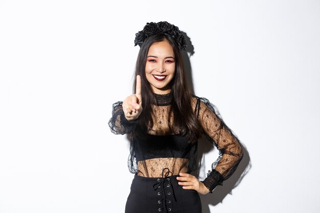Image of smiling beautiful asian woman in halloween costume showing stop gesture, extend one finger and looking happy, prohibit or reject something, standing white background.