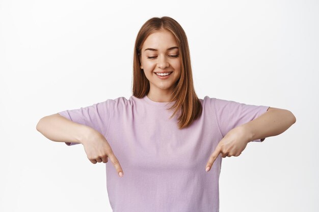 Image of smiling blond girl pointing, looking down with pleased face, makes her choice, showing advertisement, event announcement, stands against white background.