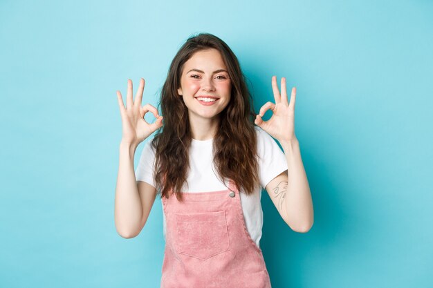 Image of smiling brunette female model say yes, showing okay signs in approval, agree or praise good choice, recommending product, standing against blue background.