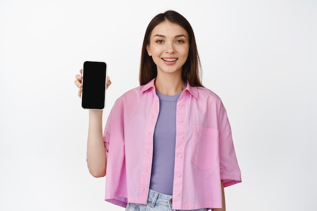 Image of smiling brunette woman showing smartphone screen recommending application demonstrating app interface white background