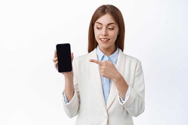 白い背景の上に立っているスマートフォンのアプリケーションを示す携帯電話の画面に指を指している企業のビジネススーツで笑顔のビジネス女性の画像