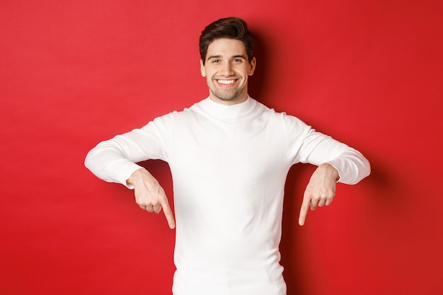 Image of smiling handsome man in white sweater inviting visit page pointing fingers down and showing...