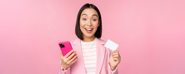 ピンクの背景に立っている携帯電話でスマートフォンアプリケーションの注文でクレジットカードをオンラインで支払うことを示す笑顔の幸せなアジアの実業家の画像