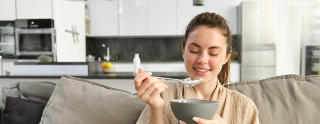 Free photo image of smiling happy young woman eating breakfast holding bowl of cereals with milk having meal at