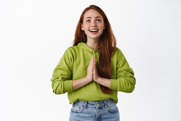 Image of smiling hopeful girl makes wish, holding hands in pray, praying or supplicating, wishing for something, looking up in sky, white background