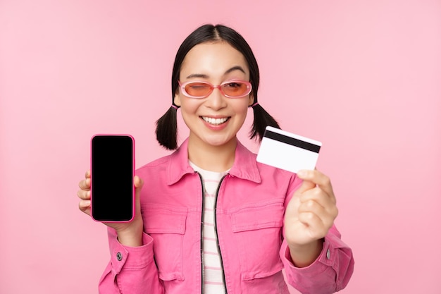 ピンクの背景の上に立ってオンラインショッピングの非接触型決済を支払うクレジットカードと携帯電話の画面のスマートフォンアプリケーションインターフェイスを示す笑顔の韓国人女性の画像