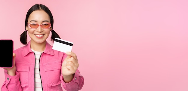 Image of smiling korean woman showing credit card and mobile phone screen smartphone application interface paying online shopping contactless standing over pink background