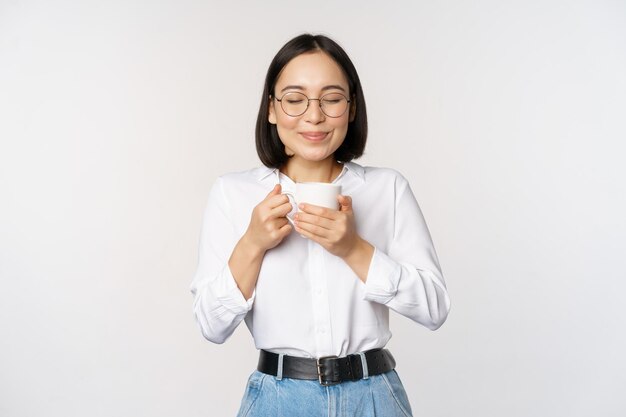 白い背景の上に立っておいしい飲み物を楽しんでマグカップで香りのコーヒーを嗅ぐ笑顔の韓国人女性の画像