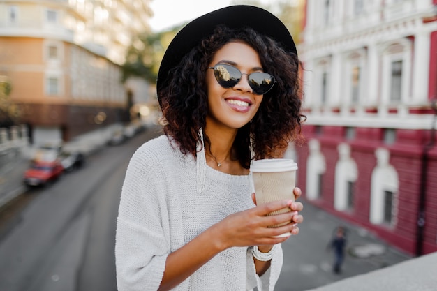 Image of smiling pretty black woman in white sweater and black hat  enjoying   coffee to go.  Urban background.