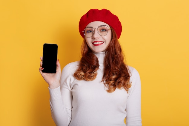 Image of smiling red haired girl wearing red beret and white shirt, girl with red lips holding smart phone with blank screen in hands on yellow